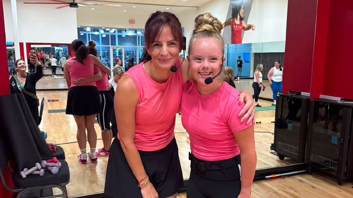 Two fitness instructors wearing headsets and pink shirts stand together in a dance studio.